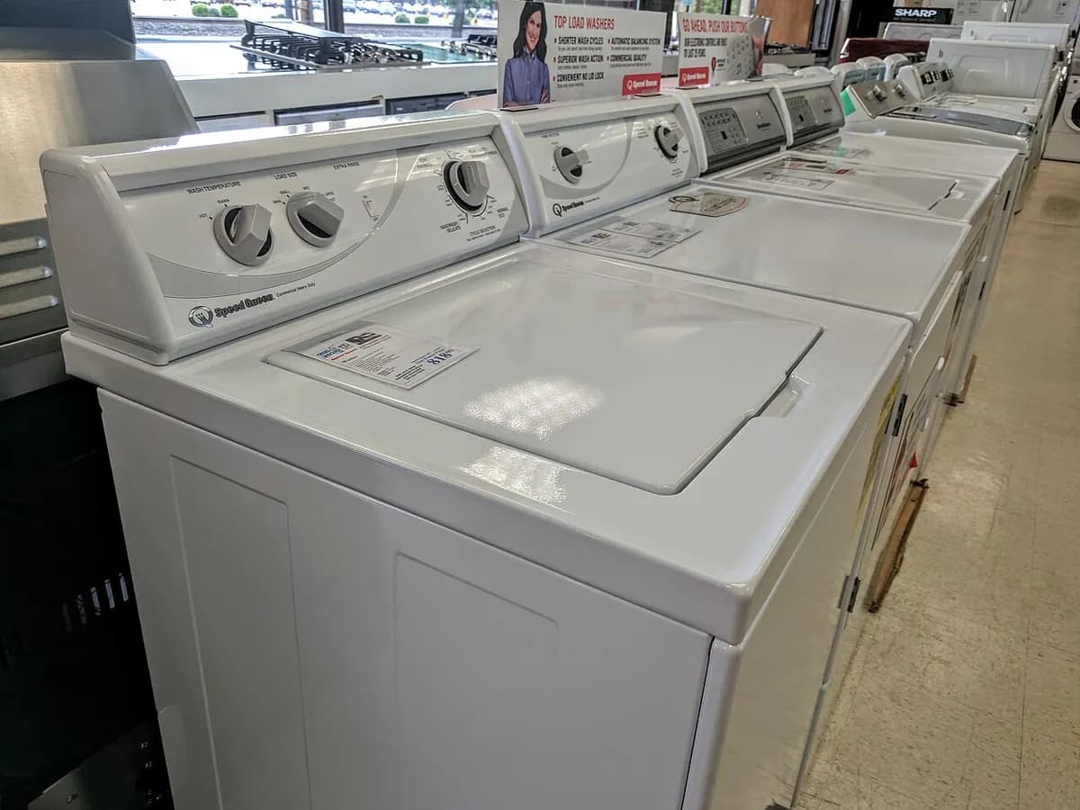 A row of white top-loading washers and dryers lined up in an appliance store.
