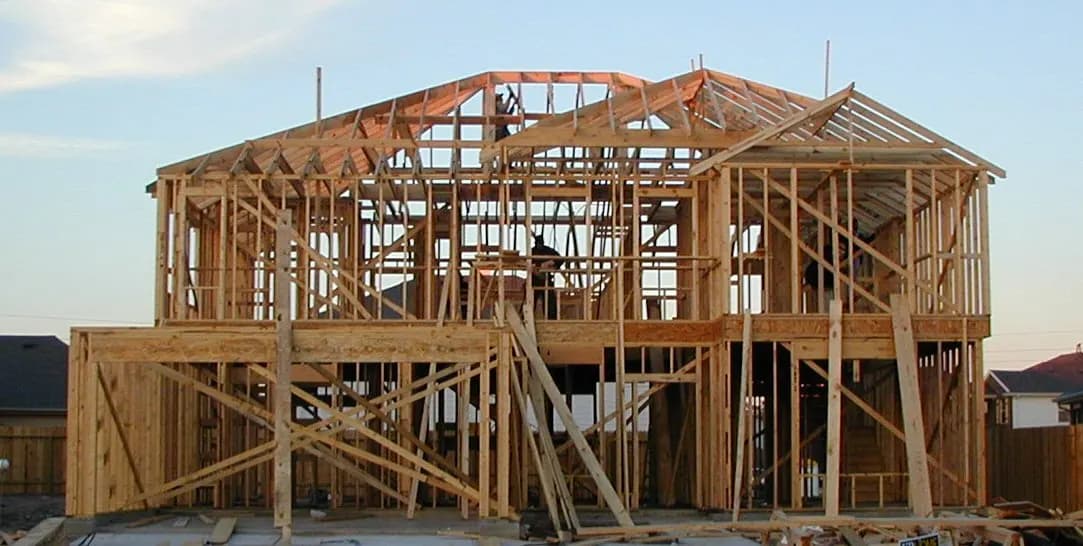 A two-story house frame of lumber studs and rafters under construction against a sky.