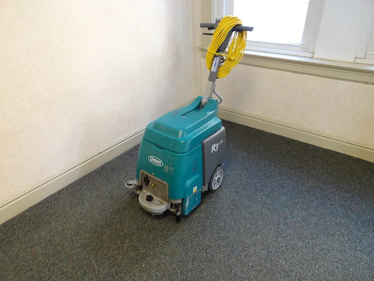 A walk-behind floor scrubbing machine with a yellow cord parked in the corner of a carpeted room.