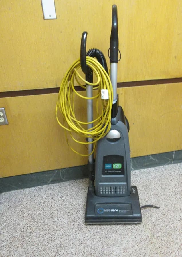 An upright vacuum cleaner with a coiled yellow cord hanging from the handle, against a yellow wall.