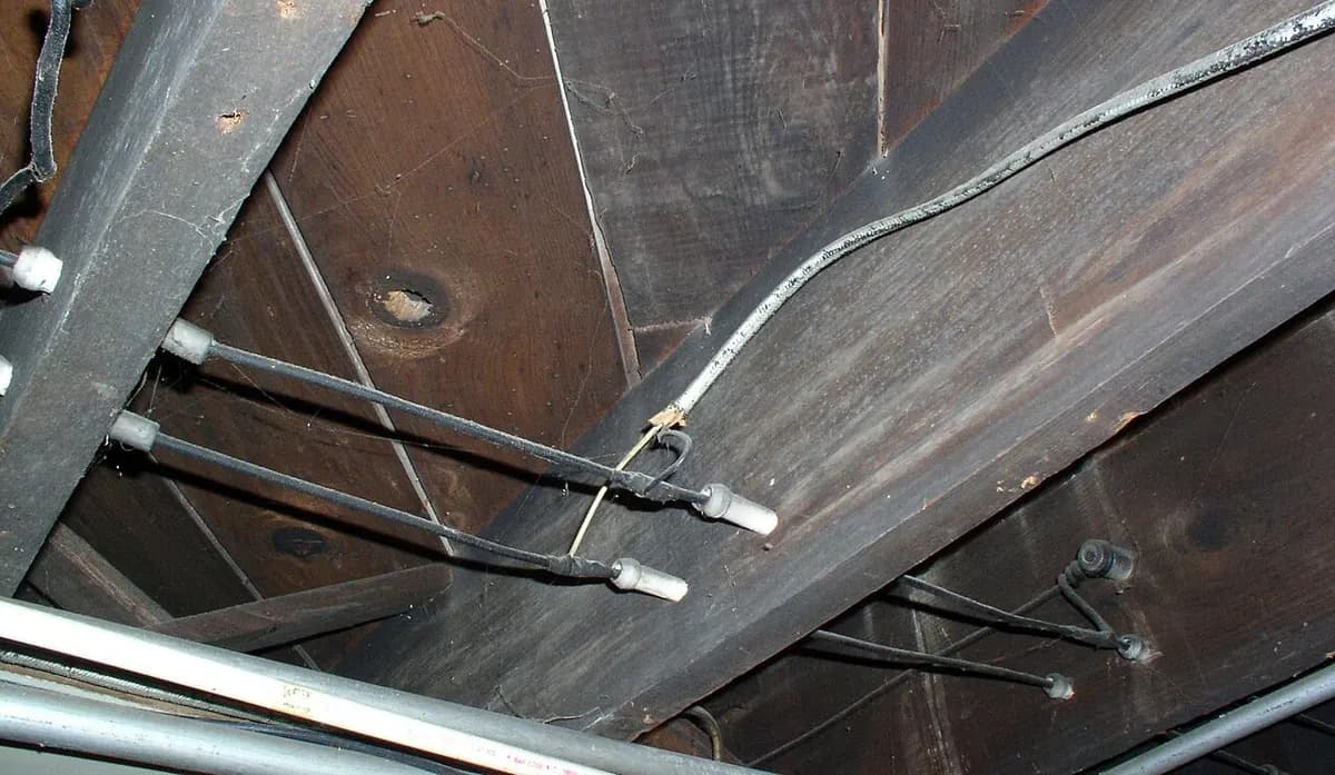Old wires running along wooden joists in a basement ceiling, held on porcelain insulators.