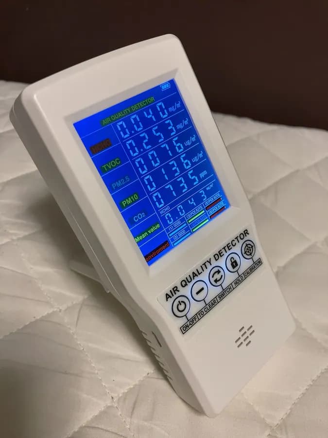 A white handheld air-quality monitor with a blue screen standing on a white surface.