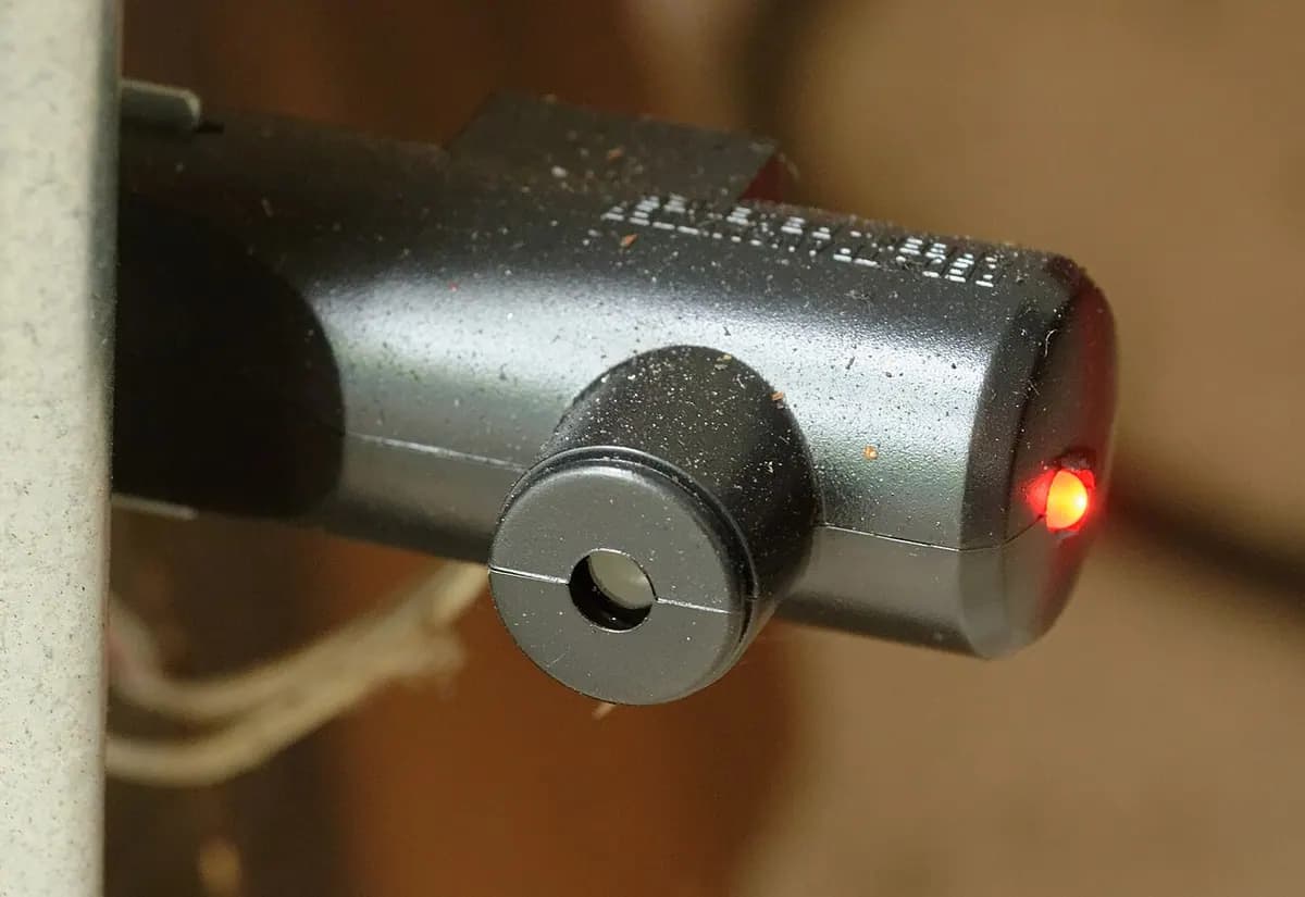 A small gray photo-eye sensor with a glowing red light, mounted near the floor beside a garage door.