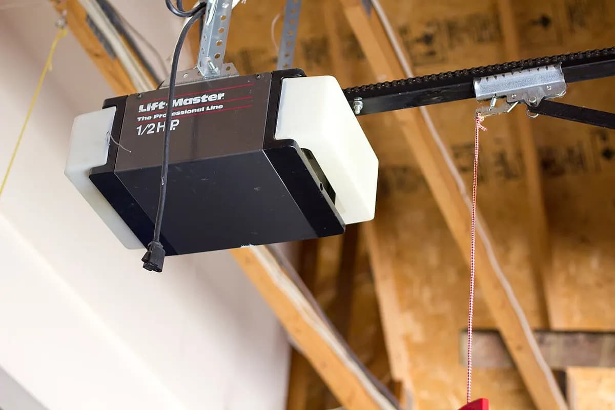 A garage door opener motor unit mounted to ceiling rafters with a rail and a release cord hanging down.