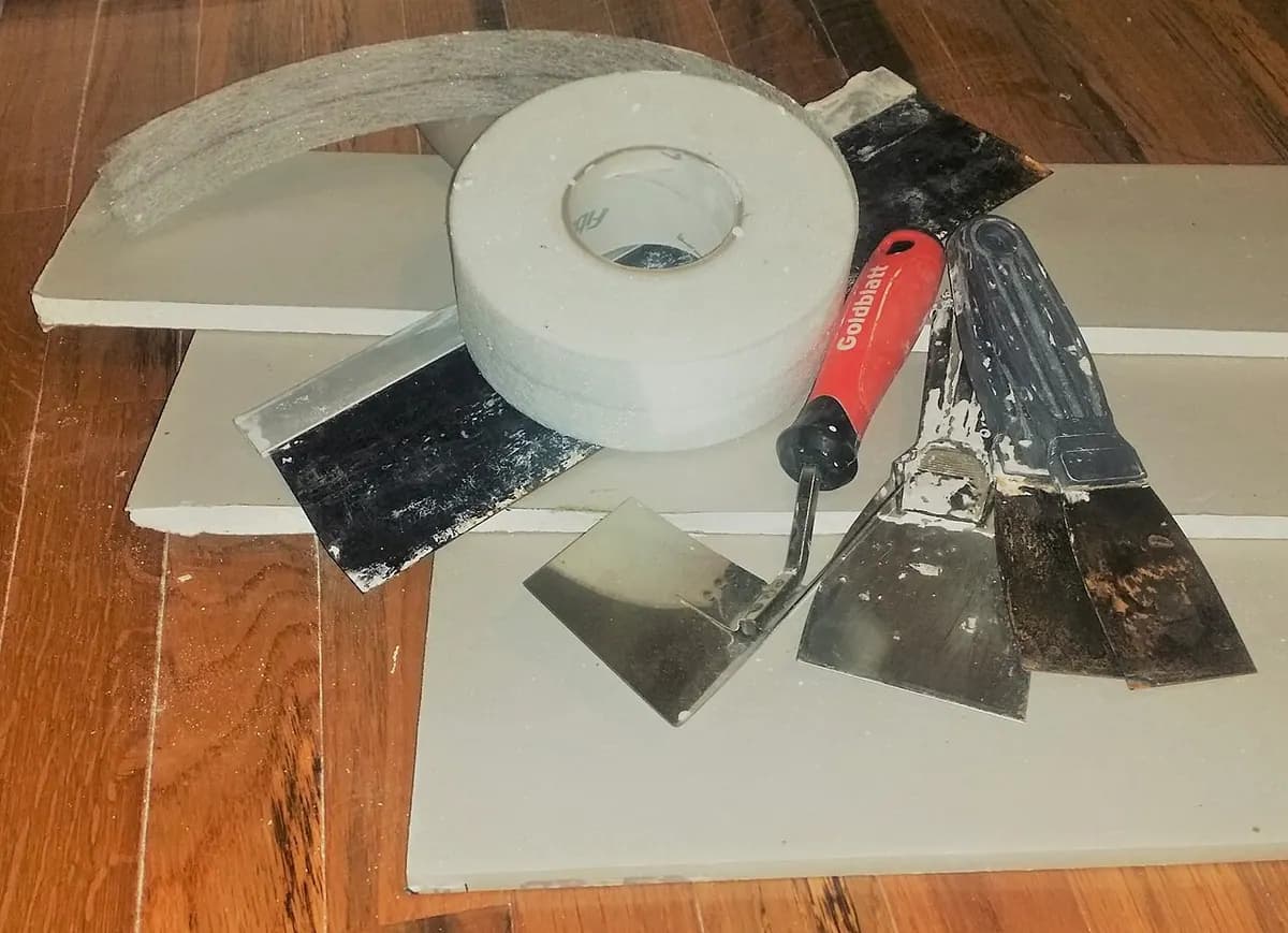 Drywall tape, putty knives and sheets of drywall laid on a wooden surface.