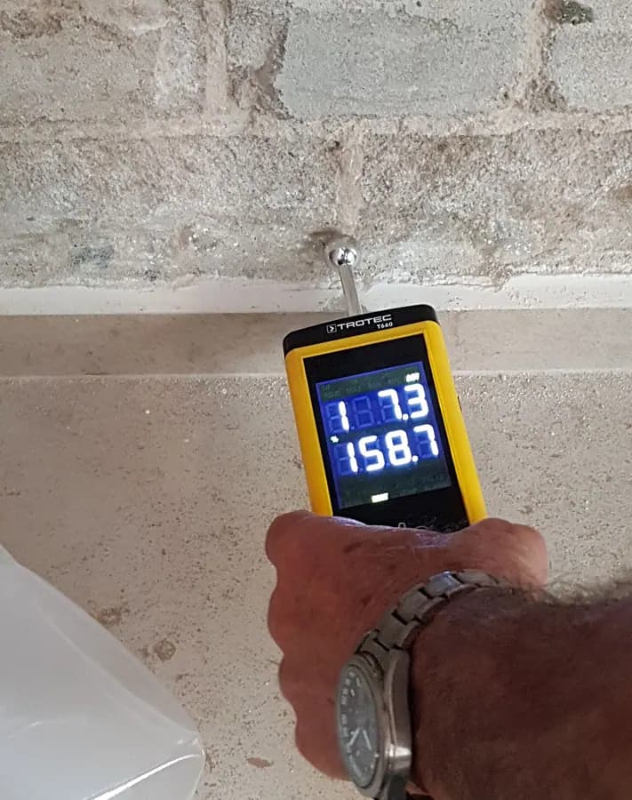 A hand holding a yellow moisture meter against a wall, with a digital reading on its screen.