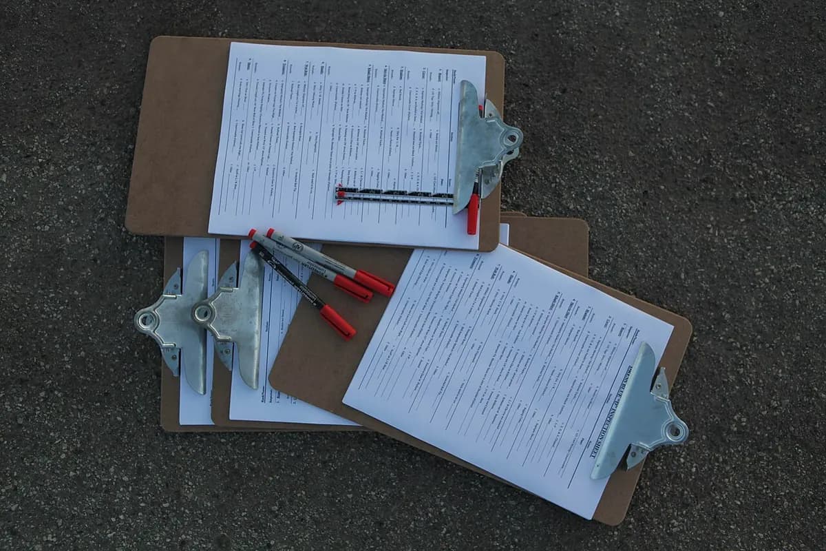 Two clipboards with printed checklists lying on dark pavement, with pens.