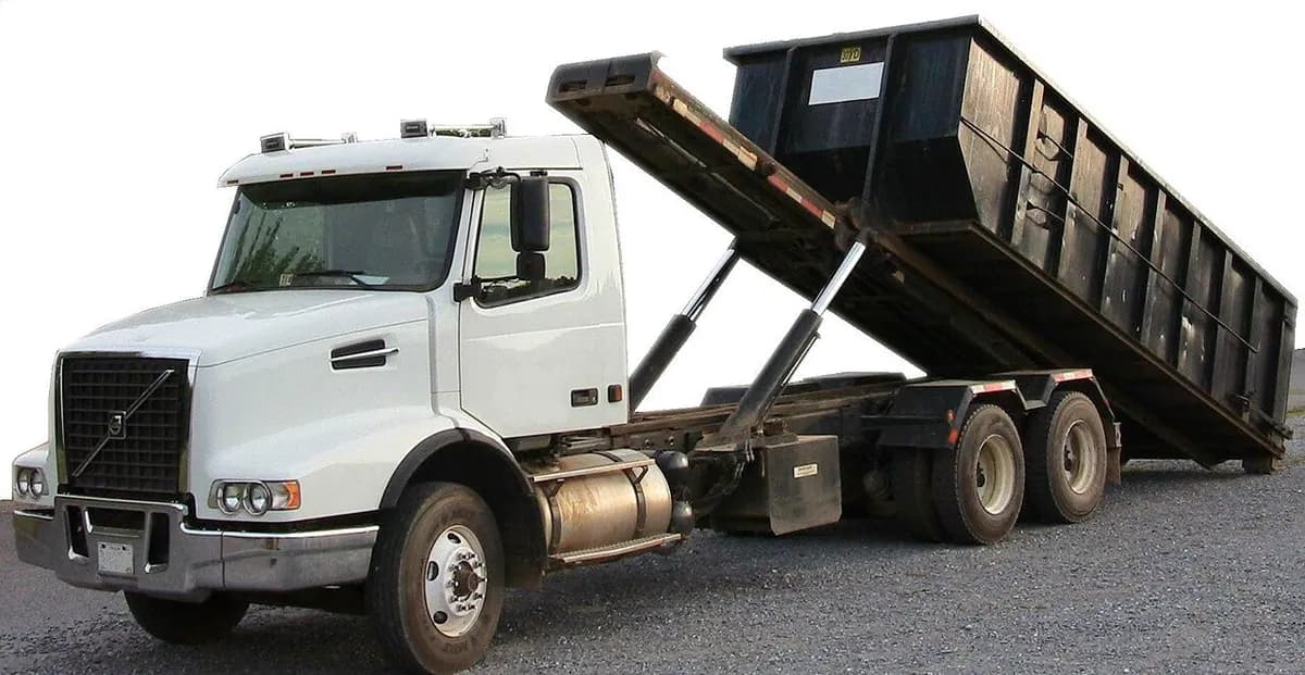 A roll-off truck tilting its tall container to unload on a paved lot.