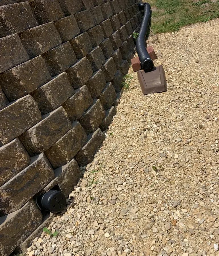 A stacked block retaining wall with gravel behind it and the end of a drain pipe showing.