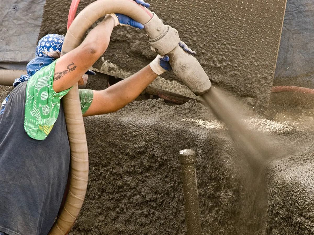 A worker's hands holding a hose that sprays concrete onto the earth walls of a pool excavation.