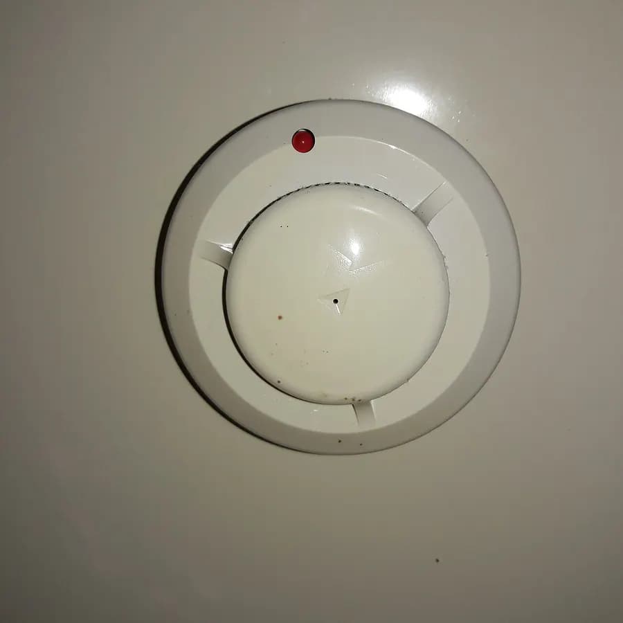 A round beige smoke alarm with a small red light, mounted on a ceiling.