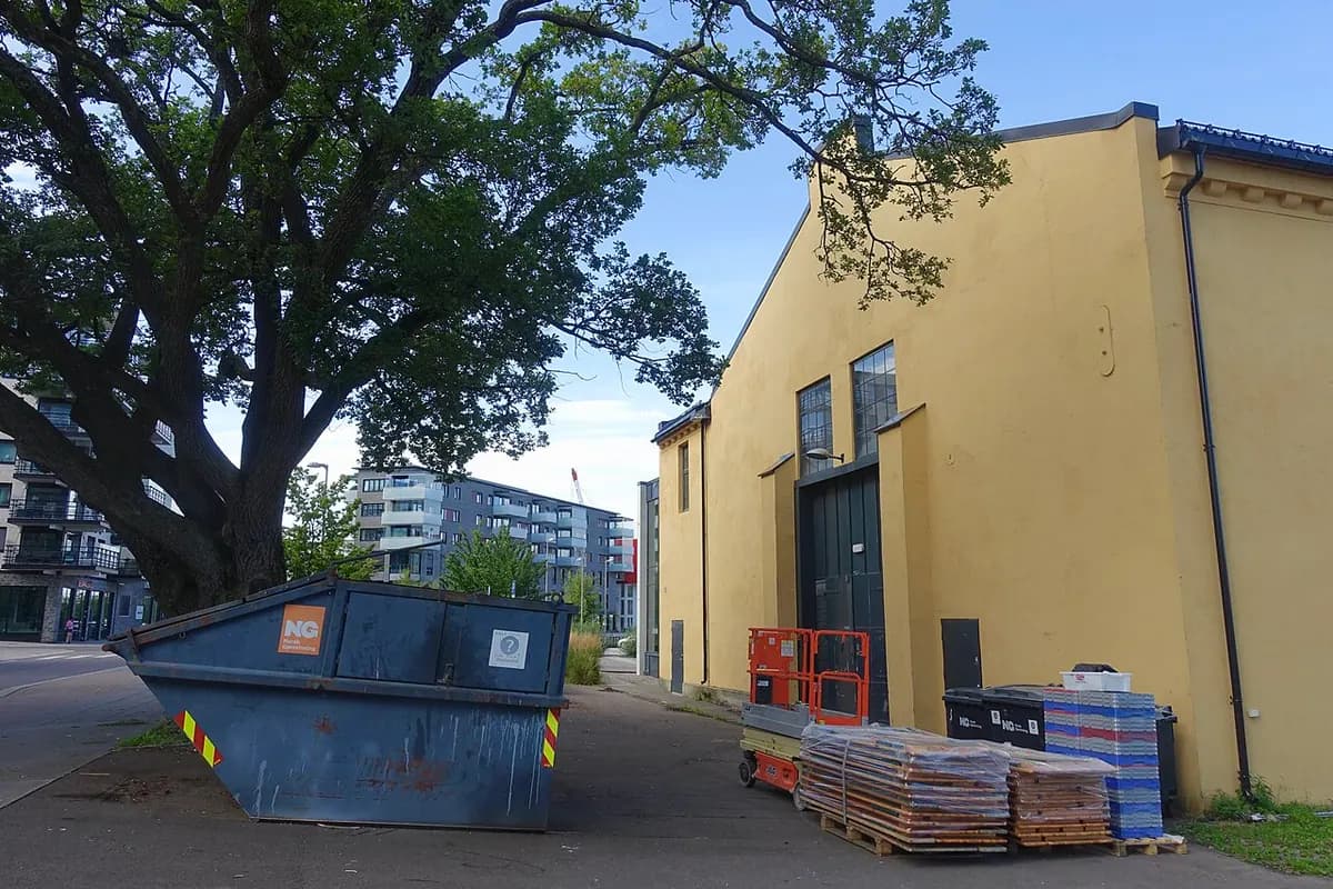 A blue dumpster beside a yellow industrial building with stacked materials next to it.