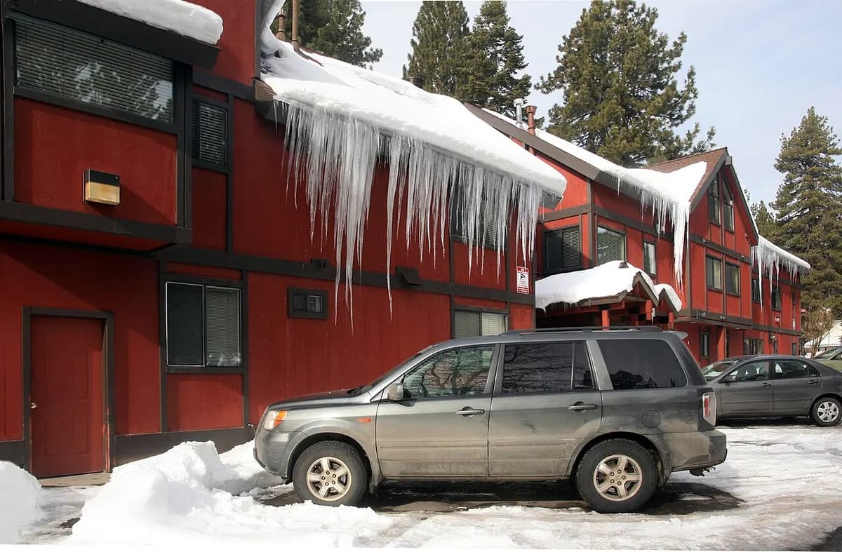 Long icicles hanging from the edge of a snowy roof above a parked car.