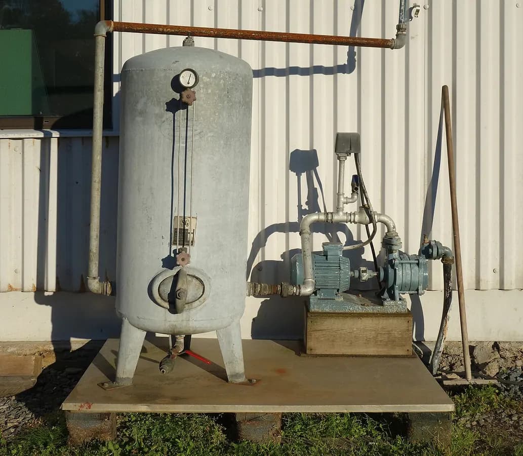 A vertical pressure tank next to a pump beside a metal-sided building.