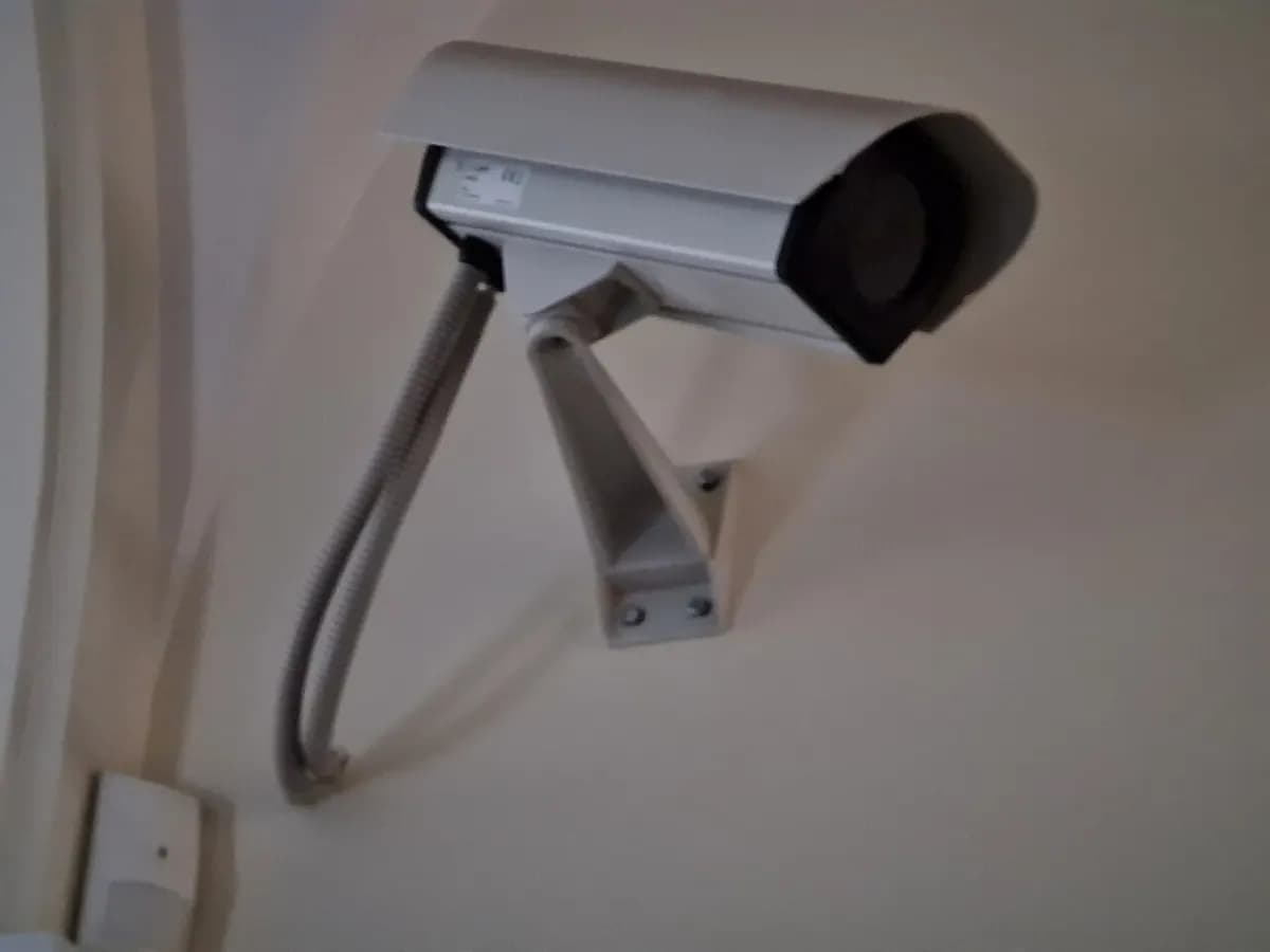 A gray security camera mounted on a wall bracket with its cable running down the wall.