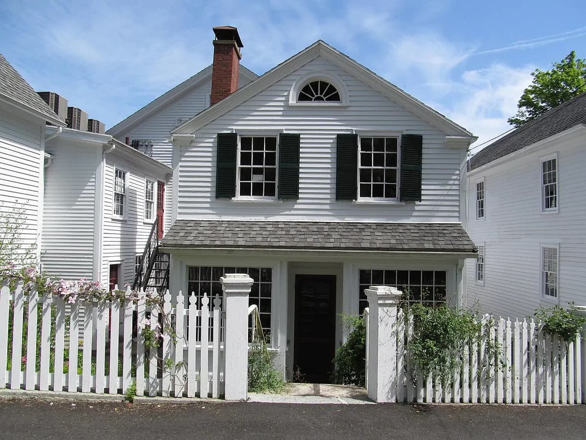 A white clapboard house with black shutters, a front door with a fan-shaped window above it and a white picket fence.