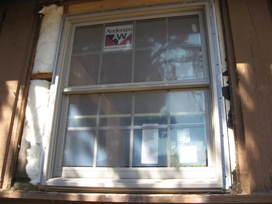 A large double-hung window with a grid pattern in an exterior wall, with old trim and insulation visible around the frame.